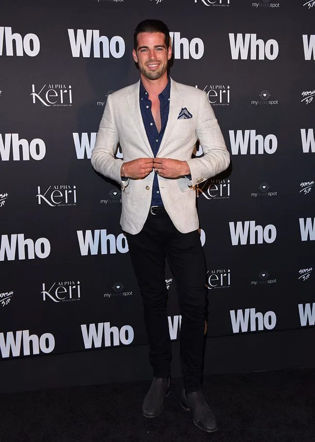 Man in a light blazer and dark pants posing at a "Who" event backdrop.