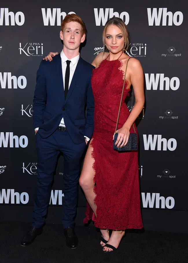Young man in a blue suit and woman in a red dress pose on the red carpet at the "Who" event.