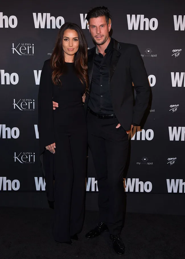 A couple poses at the "Who" magazine event, dressed in black, with a branded backdrop behind them.