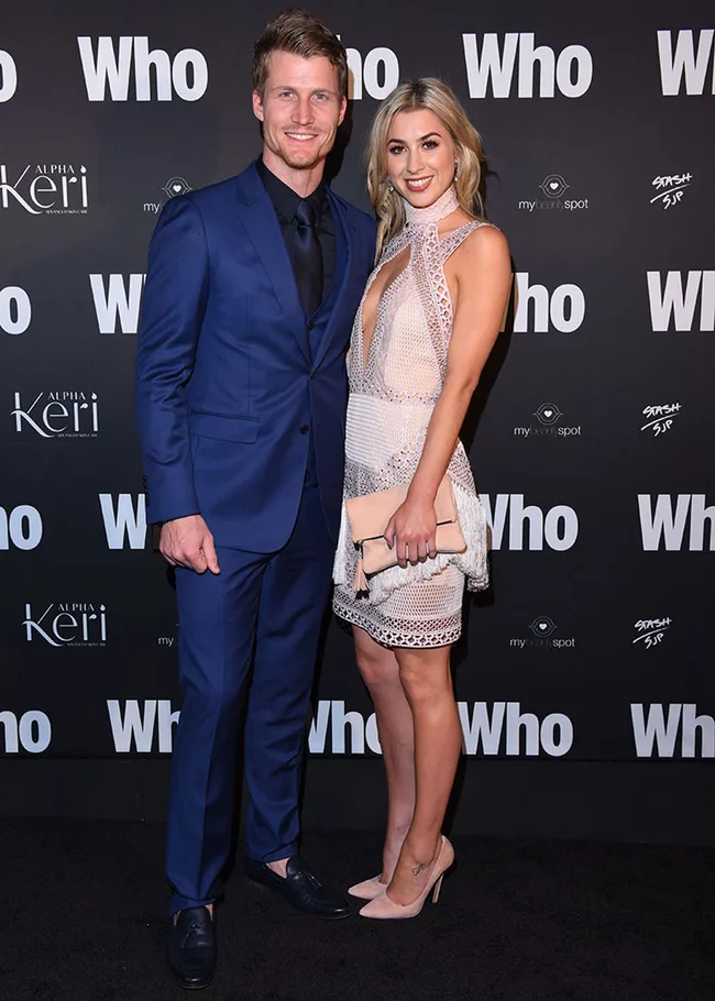 A man in a blue suit and a woman in a white dress pose on the red carpet at a "Who" magazine event.