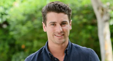 Man in a dark shirt smiling outdoors with blurred green background.