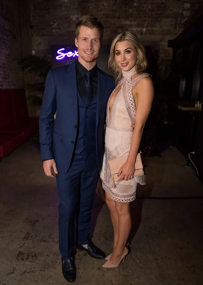 A man in a blue suit and a woman in a mesh dress pose together at an event with "Sex" sign in background.