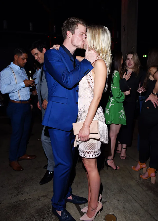 A couple kisses in a lively party setting with guests mingling in the background.