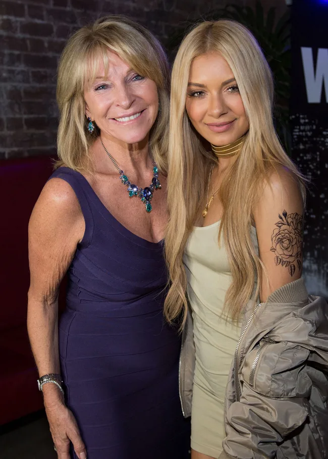 Two women smiling and posing together, one with a floral tattoo on her arm, indoors.