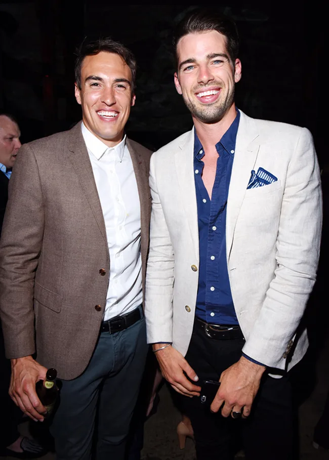Two men in blazers smiling at a party. One holds a bottle, the other has his hand in his pocket.