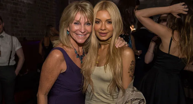 Two women smiling and posing together at a social event, one with long blonde hair and the other with short blonde hair.