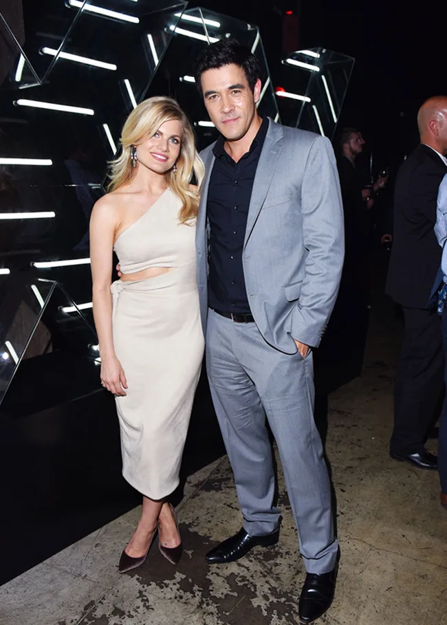 A woman in a white dress and a man in a gray suit pose together at an event with neon lights in the background.