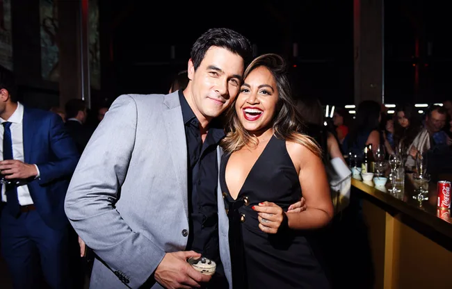 Two people smiling and posing together at an indoor event, with others socializing in the background.