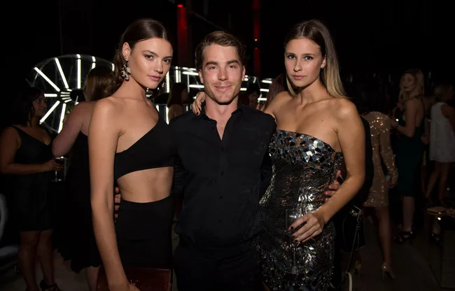 Three people posing together at an event, two women and a man, dressed in evening wear with a dark, ambient background.