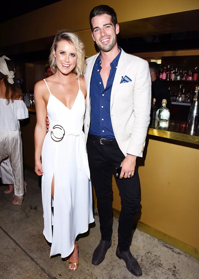A woman in a white dress and a man in a light blazer stand smiling at a bar.