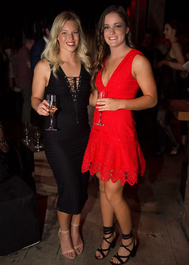 Two women in black and red dresses holding champagne glasses at a social event, smiling at the camera.