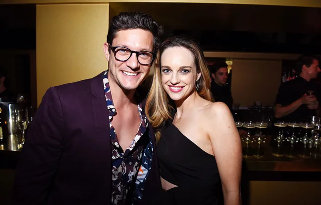 Two people smiling and posing in a bar setting at an event, with drinks visible in the background.