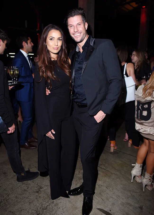 A couple in formal black attire smiles at a social event, surrounded by other guests.
