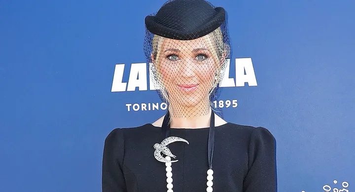 Woman in black dress with bird brooch, wearing a hat with netting, standing in front of blue Lavazza backdrop.