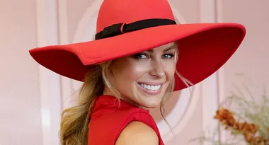 A woman smiling, wearing a large red hat with a black band and matching red outfit, poses in a stylish setting.