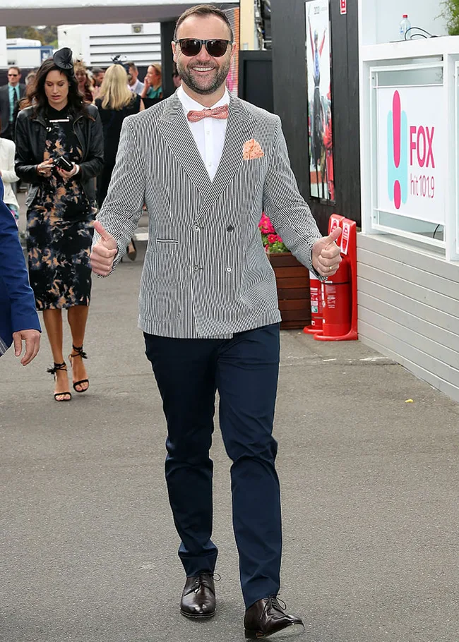 Man in striped blazer, navy pants, and sunglasses giving a thumbs-up while walking outdoors.