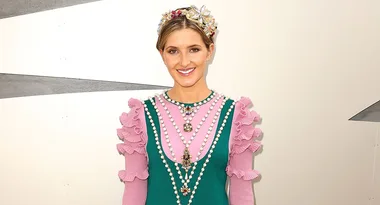 A smiling woman wearing a pink and green ruffled dress with pearl details and a decorative headpiece.