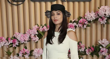 Woman in white dress and black hat posing in front of floral backdrop with pink flowers.