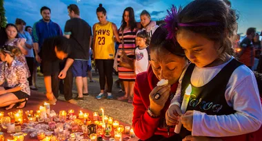 A vigil with adults and children holding candles, surrounded by lit candles on the ground during dusk.