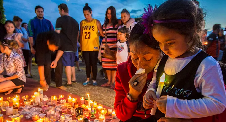 A vigil with adults and children holding candles, surrounded by lit candles on the ground during dusk.