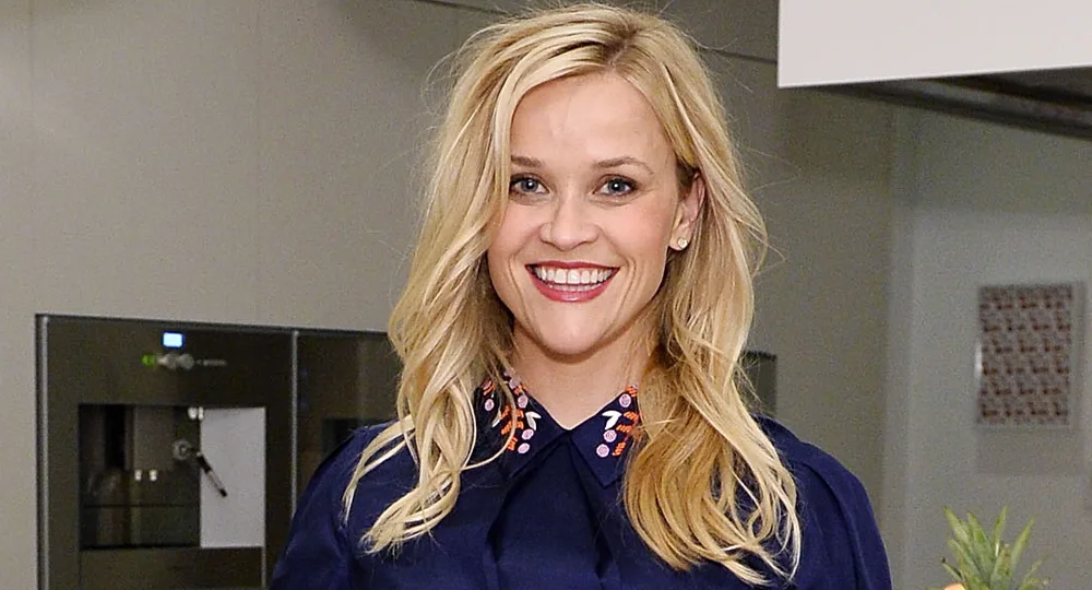 A smiling woman with long blonde hair, wearing a navy top with a decorative collar, in a kitchen setting.