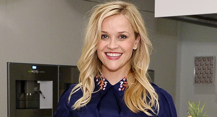 A smiling woman with long blonde hair, wearing a navy top with a decorative collar, in a kitchen setting.