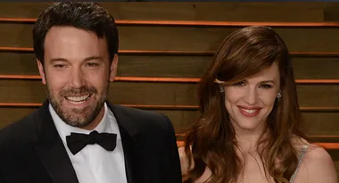 A man in a black tuxedo and a woman in a light-colored gown smile together at a formal event.