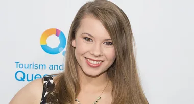 A person smiling at an event, with a "Tourism and Queensland" backdrop.