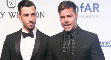 Two men in tuxedos pose for a photo at an event with an amfAR backdrop.