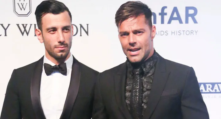 Two men in tuxedos pose for a photo at an event with an amfAR backdrop.