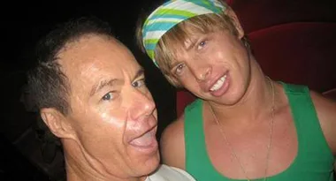 Two people smiling, one wearing a green tank top and headband, taking a selfie against a dark background.