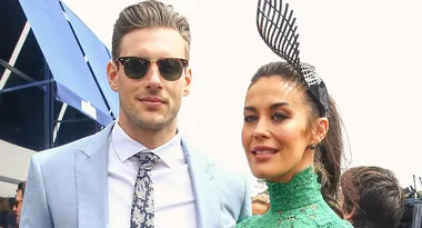 Two people at an outdoor event; man in a suit with sunglasses, woman in green dress with decorative headpiece.
