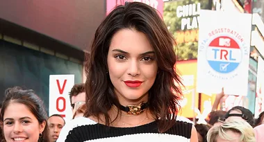 ```html
<img src="image.jpg" alt="Person with dark hair and red lipstick, standing outdoors with a crowd and signs in the background." />
```