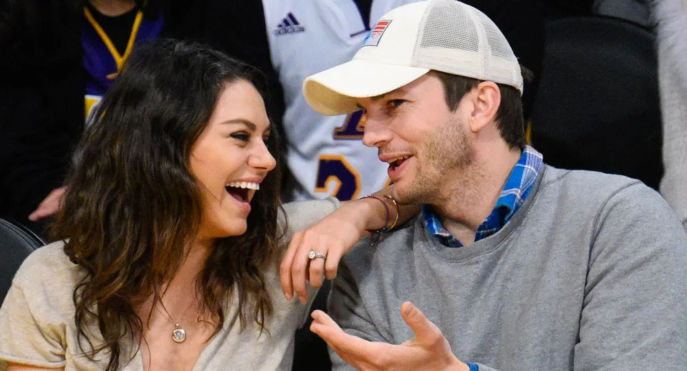 A man and a woman, both smiling and engaged in conversation, seated at what appears to be a sports event.
