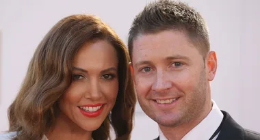 Two people smiling at the camera, one with long brown hair and red lipstick, the other with short hair in a suit.