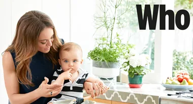 A woman and a baby in a kitchen, with the word "Who" on the wall; plants and fruits are on the counter.