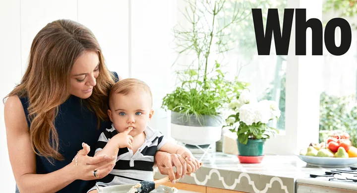 A woman and a baby in a kitchen, with the word "Who" on the wall; plants and fruits are on the counter.