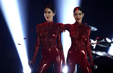 Two performers in glittery red outfits stand on stage with spotlights behind them.