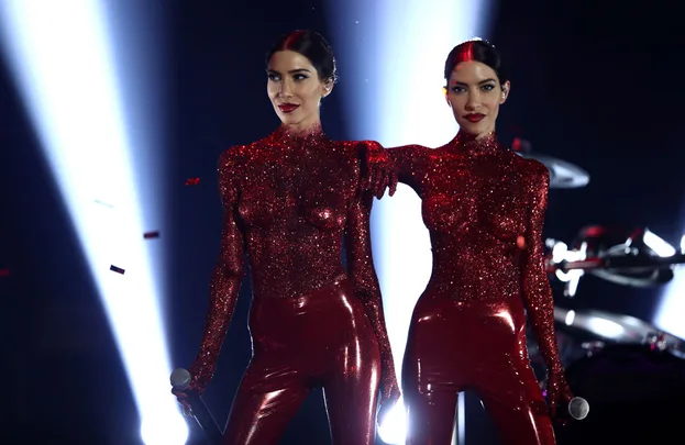 Two performers in glittery red outfits stand on stage with spotlights behind them.