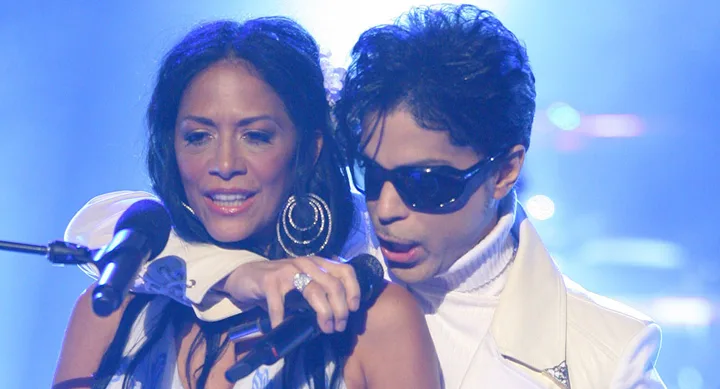 Two performers on stage, one wearing sunglasses, singing into microphones with a blue-lit background.