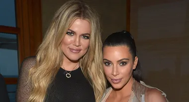 Two women pose together indoors, one with long blonde hair, the other with dark hair tied back, both smiling at the camera.