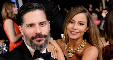 Couple smiling at an event, with a woman wearing a gold necklace and a man in a suit with a black beard.