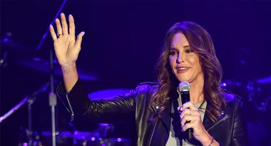 Caitlyn Jenner speaking on stage, holding a microphone and raising her hand, wearing a leather jacket.