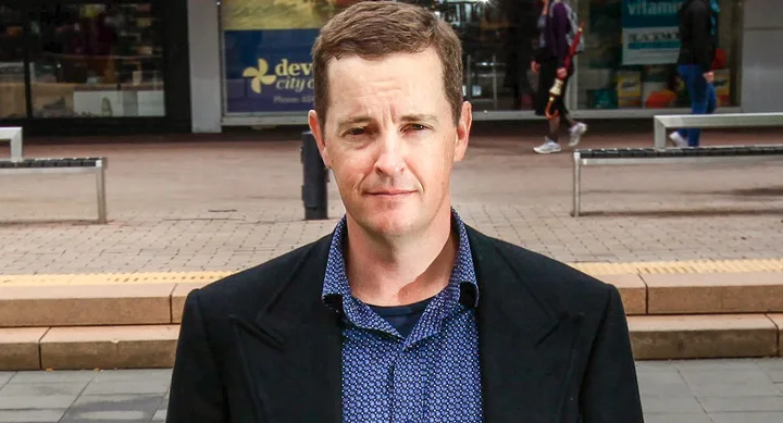 Man in a blue shirt and dark jacket standing on a city street with benches and shops in the background.
