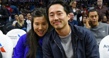 A couple smiling at a sports event, seated with people in the background.