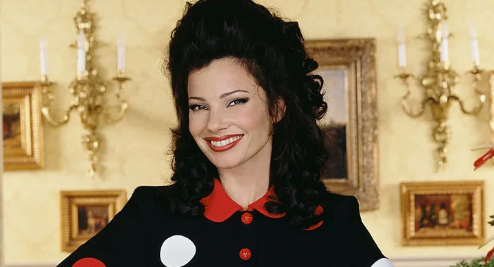 A smiling woman with dark hair in a black and red outfit stands in a room with ornate decor and paintings.