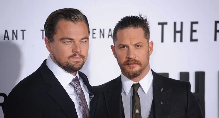 Two men in suits at a movie premiere with "The Revenant" in the background.