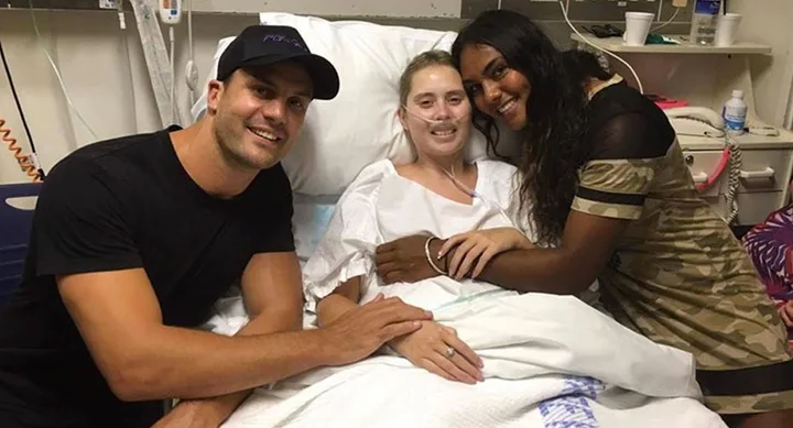 Three people smile in a hospital room; one is in a bed, and two are standing beside them.