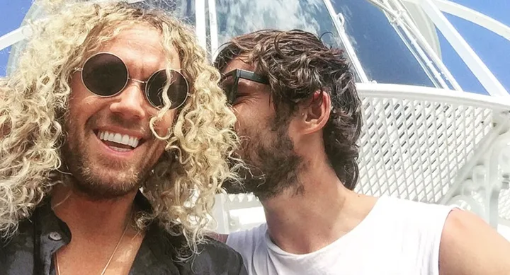 Two men smiling outdoors, one with curly hair and round sunglasses, the other whispering with short hair and beard.