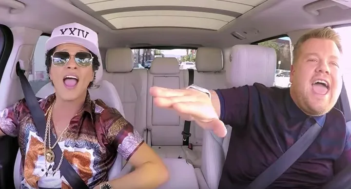 Two people singing enthusiastically inside a moving car, one wearing sunglasses and a cap with "XXIV" on it.